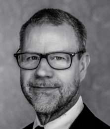 Man med glasögon och skägg ler i svartvitt porträtt.