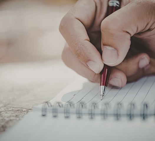 A hand holds a red pen, writing on a spiral-bound notebook with lined paper.